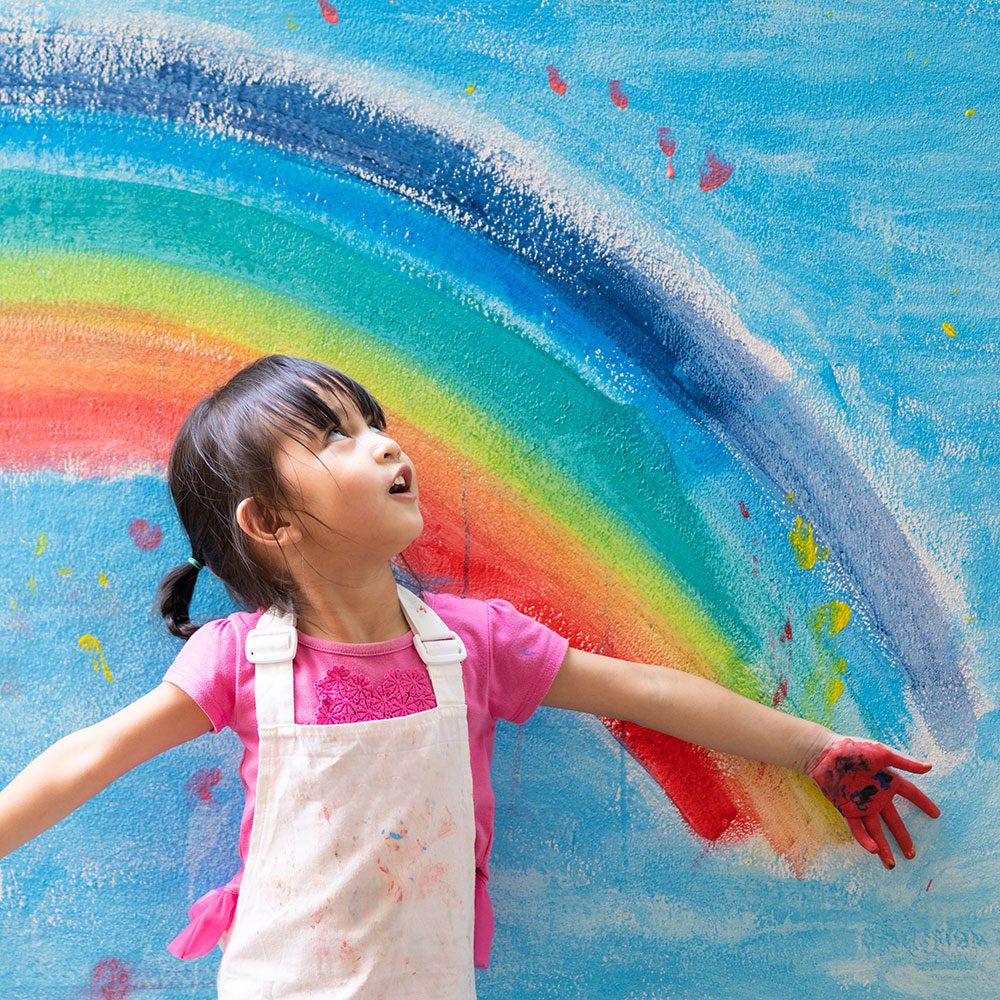 A joyful girl in front of a colourful rainbow on a wall.