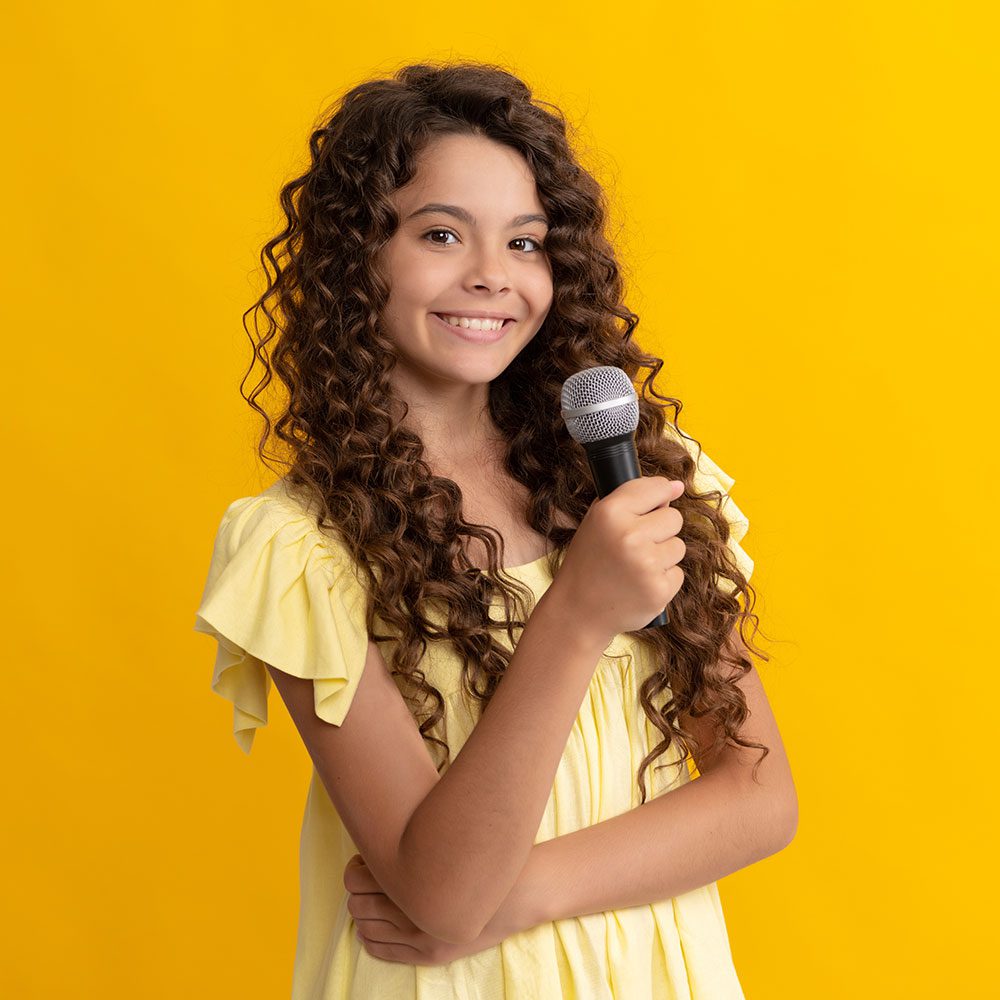 A young girl confidently holds a microphone, ready to captivate the audience, against a vibrant yellow background.