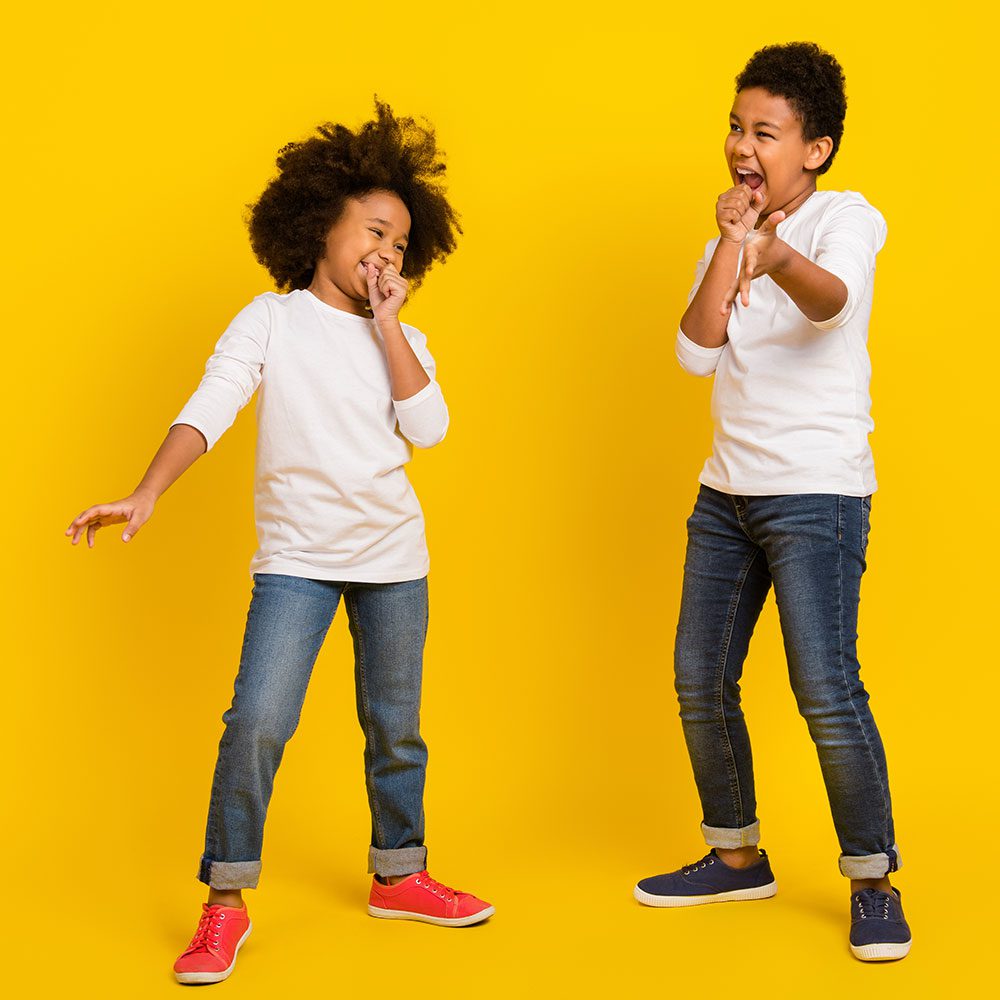 Two kids in white shirts and jeans posing on a yellow background.
